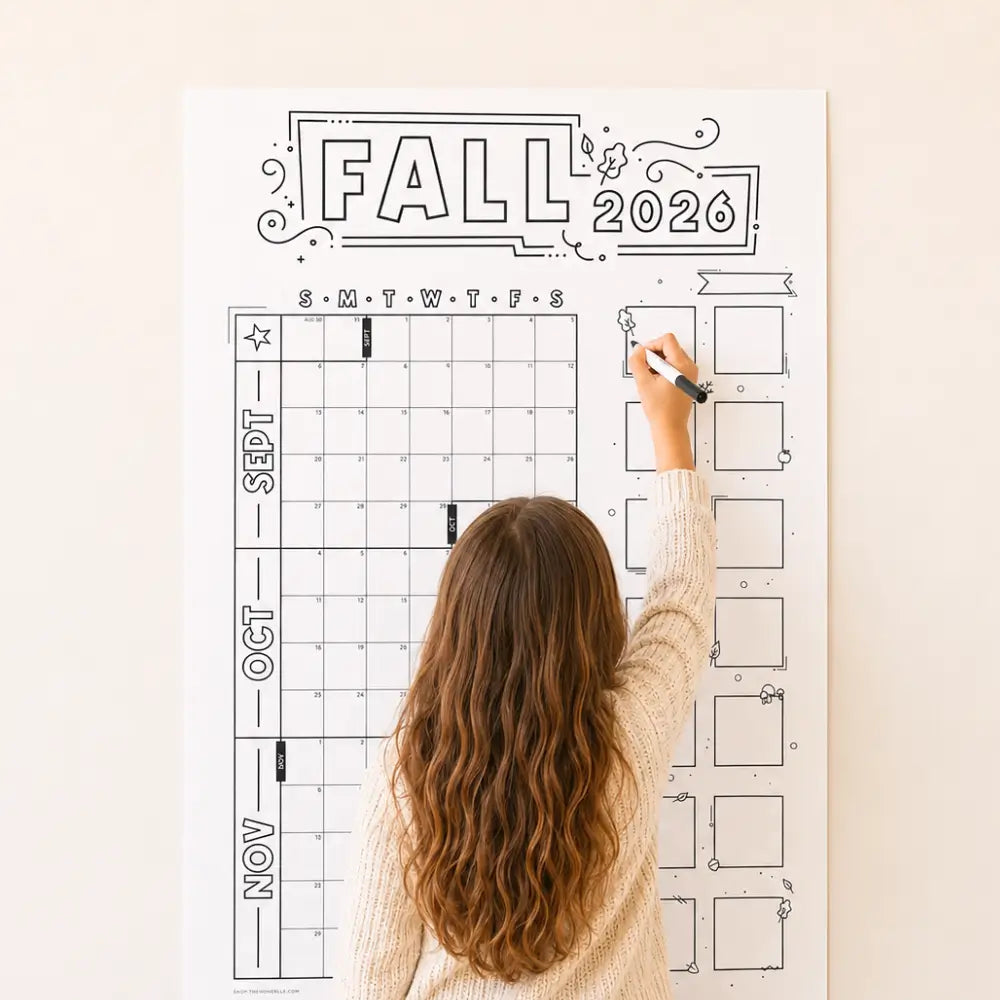 A girl with long wavy brown hair, seen from behind, writing on a large white Fall 2026 planner calendar poster mounted on a light beige wall.