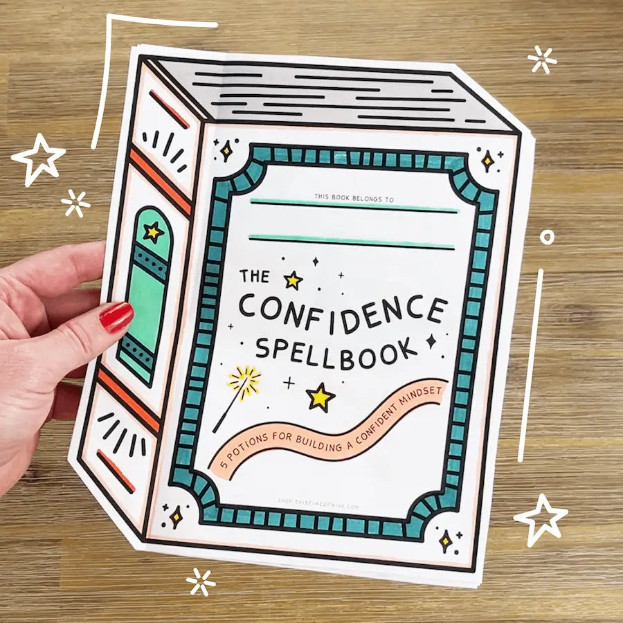 Hand holding a 'The Confidence Spellbook' book-shaped card on a wooden background